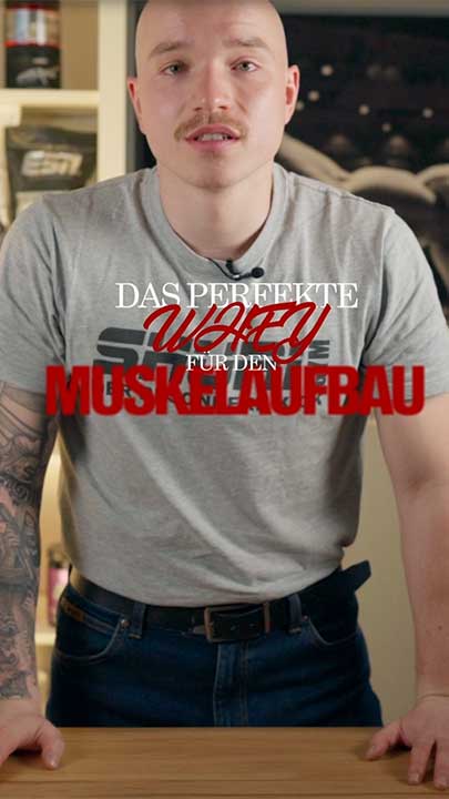 Nico von Kraftmahl steht an einem Tisch, trĂ€gt ein graues T-Shirt mit Logo â Text eingeblendet: âDas perfekte Whey fĂŒr den Muskelaufbauâ.
