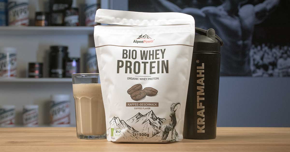AlpenPower Bio Whey Protein im Kaffeegeschmack neben Kraftmahl Shaker und Glas Proteinshake auf Holztisch.