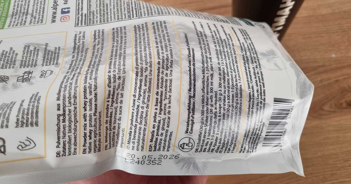 AlpenPower Whey Isolat Vanille – Rückseite der Verpackung mit Chargennummer und Mindesthaltbarkeitsdatum (20.05.2026), gehalten in einer Hand auf Holztisch – als Beleg für eigenen Produkttest