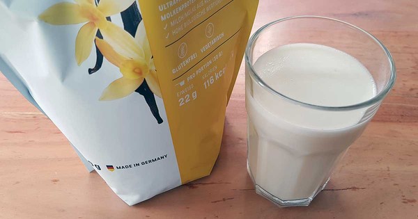 BetterProtein Whey Eiweißpulver neben einem frisch zubereiteten Proteinshake im Glas auf Holztisch