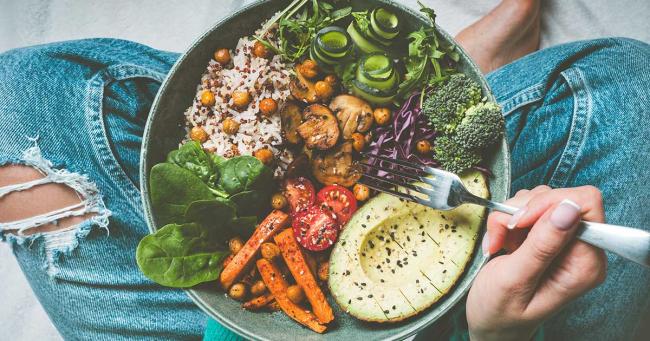 Bunte vegane Bowl mit Avocado, Reis, Kichererbsen, Spinat, Brokkoli, Karotten, Gurke, Tomaten und Pilzen auf dem Schoß einer Person in Jeans