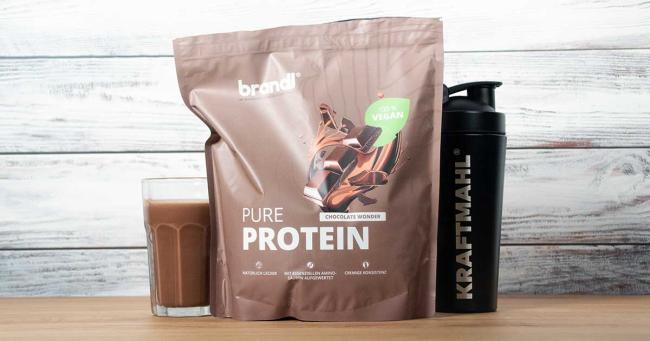 Brandl Pure Protein Vegan 2.0 in der Geschmacksrichtung Chocolate Wonder – neben einem Glas Shake und einem schwarzen Kraftmahl-Shaker