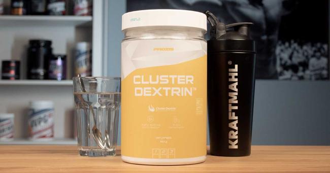 Prozis Cluster Dextrin Pulver in Dose auf Holztisch, daneben Glas Wasser und schwarzer Kraftmahl-Shaker
