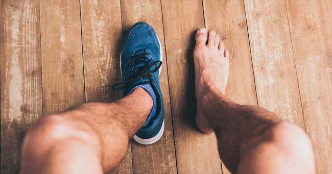 Sportler steht auf Holzboden mit einem Sneaker und einem barfußen Fuß – Vergleich von Schuh und nacktem Fuß im Trainingsalltag