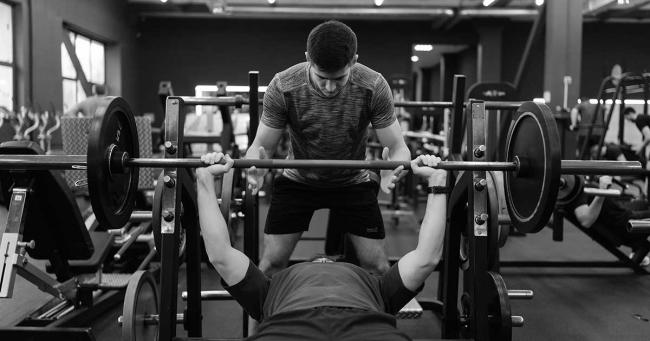 Kontrolliertes Bankdrücken mit Spotter im Fitnessstudio als Beispiel für saubere Technik und sinnvolle Progression im Krafttraining.