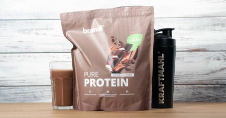 Brandl Pure Protein Vegan 2.0 in der Geschmacksrichtung Chocolate Wonder – neben einem Glas Shake und einem schwarzen Kraftmahl-Shaker