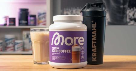 More Protein Iced Coffee im Test – Pulverdose, fertiger Drink im Glas und KRAFTMAHL Shaker auf einem Holztisch