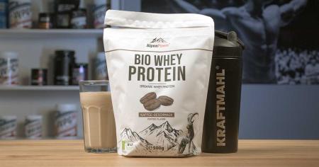 AlpenPower Bio Whey Protein im Kaffeegeschmack neben Kraftmahl Shaker und Glas Proteinshake auf Holztisch.